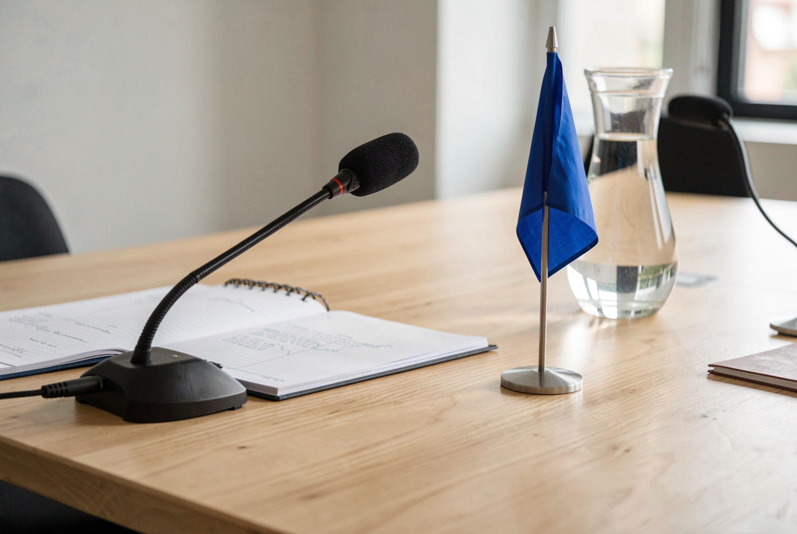 Vue rapprochée d'une table de conférence en chêne clair, sous une lumière naturelle latérale, avec un micro de délégué, un bloc-notes ouvert et un petit drapeau bleu posé près d'une carafe d'eau, cadrage serré à hauteur de table.